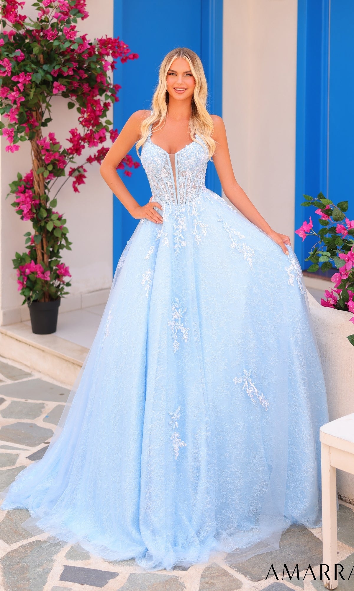 A woman stands outdoors in the Amarra 88219 Lace Prom Ball Gown, a light blue A-line dress with floral lace details. Pink flowers and blue shutters in the background enhance the romantic style of her gown.
